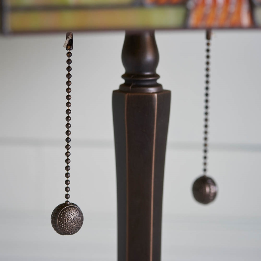 Stolová lampa s bronzovou nohou a tienidlom má dva kovové šnúrky s guľôčkami na zapínanie a vypínanie. Jeden z guľôčok má vyrytý nečitateľný text. Lampa je umiestnená na svetlom pozadí.