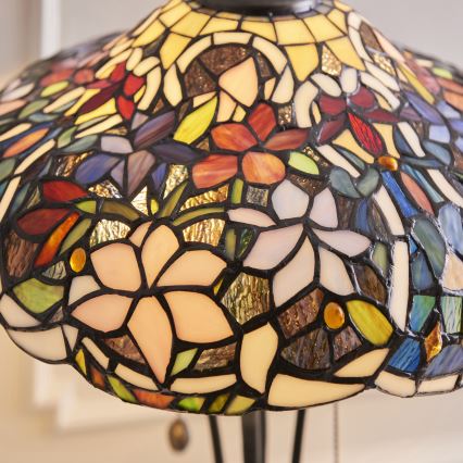 Stolová lampa s tienidlom z farebného vitráža zobrazuje rozkvitnuté kvety. Tienidlo je tvorené mnohými malými farebnými sklenenými dielikmi, ktoré sú spojené do kvetinových motívov. Lampa stojí na tmavom podstavci.
