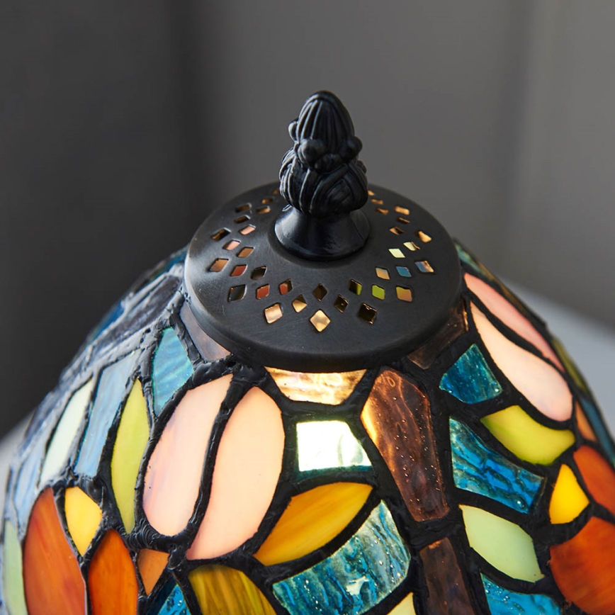 Stolová lampa svieti. Jej tienidlo je z farebného vitráža v odtieňoch ružovej, oranžovej, modrej a zelenej, s tmavo hnedými líniami. Vrchná časť lampy je čierna s otvormi.