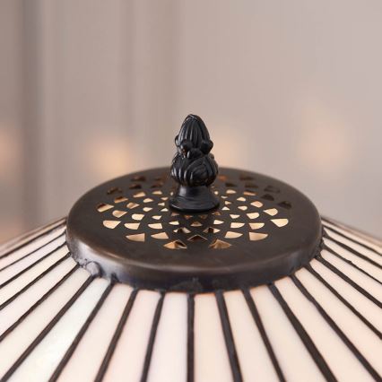 Tmavý kovový vrchlík s ozdobným vrcholom je umiestnený na tienidle stolovej lampy. Tienidlo má tvar kužeľa s vertikálnymi pruhmi.