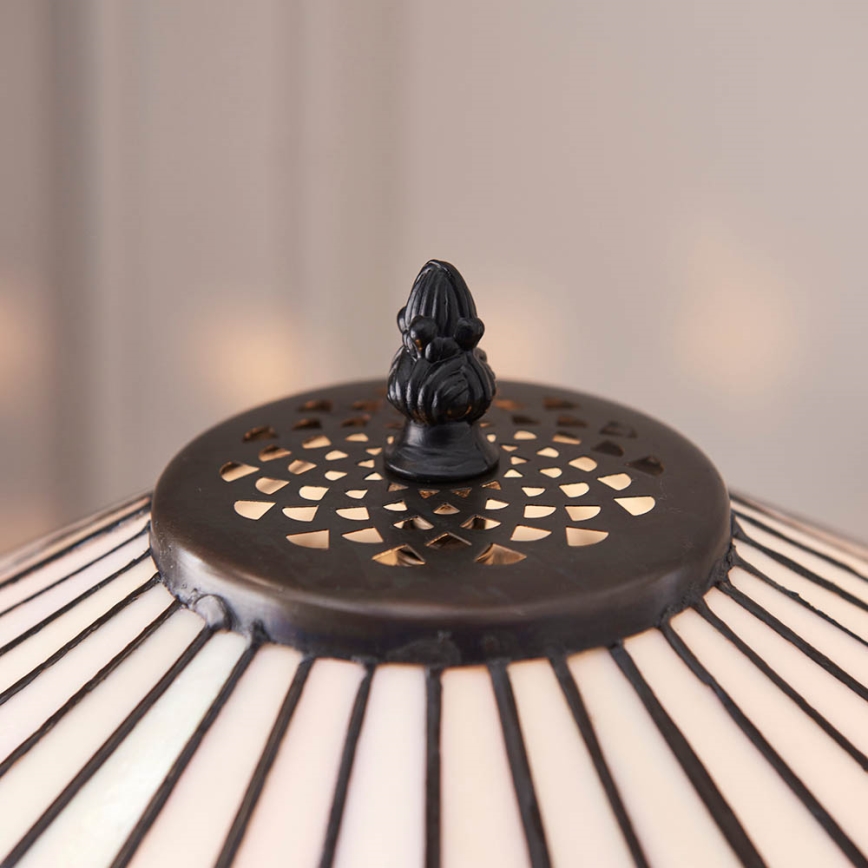 Tmavý kovový vrchlík s ozdobným vrcholom je umiestnený na tienidle stolovej lampy. Tienidlo má tvar kužeľa s vertikálnymi pruhmi.