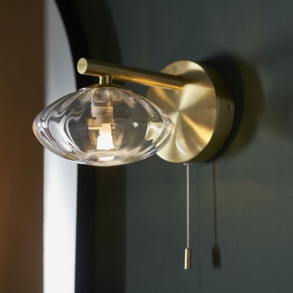 Zlatá nástenná lampa so skleneným tienidlom svieti. Lampa je pripevnená na tmavozelenej stene. Zapína sa šnúrkou.