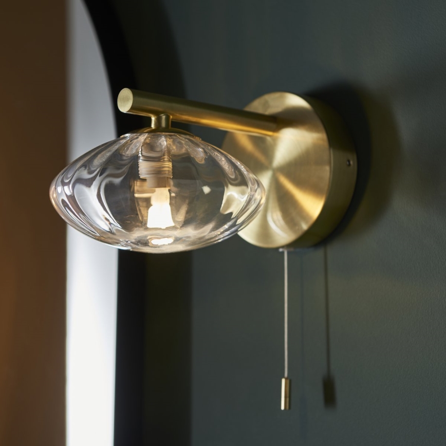Zlatá nástenná lampa so skleneným tienidlom svieti. Lampa je pripevnená na tmavozelenej stene. Zapína sa šnúrkou.