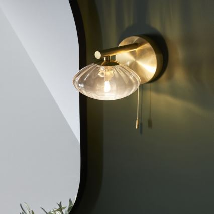 Zlatá nástenná lampa s priehľadným skleneným tienidlom v tvare mušle svieti. Lampa je pripevnená na tmavozelenej stene, v pozadí je vidieť zrkadlo. Lampa má šnúrkový vypínač.