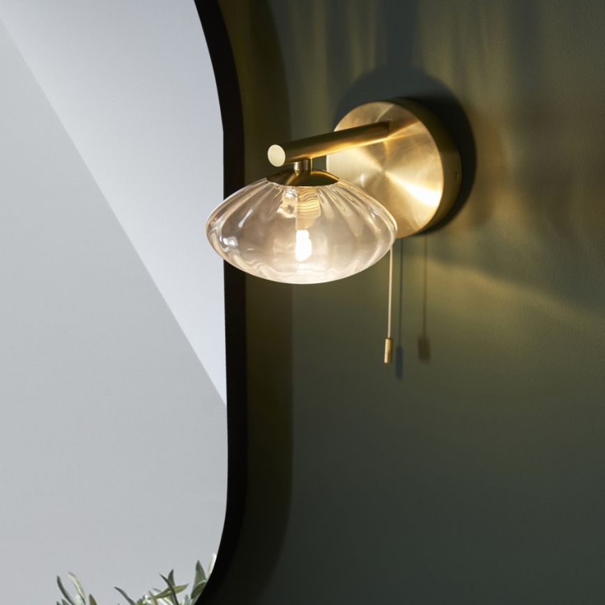 Zlatá nástenná lampa s priehľadným skleneným tienidlom v tvare mušle svieti. Lampa je pripevnená na tmavozelenej stene, v pozadí je vidieť zrkadlo. Lampa má šnúrkový vypínač.