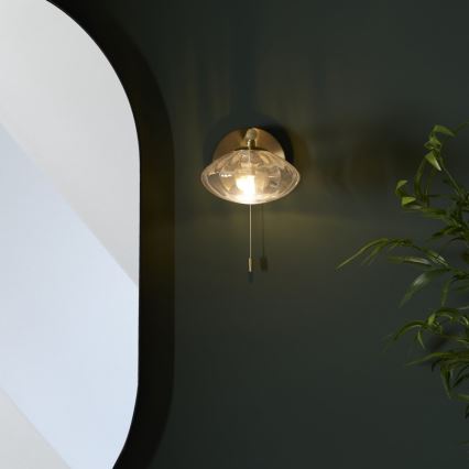 Nástenná lampa s priesvitným skleneným tienidlom svieti. Lampa je pripevnená k tmavozelenej stene, vedľa nej je časť bielej steny a zelená rastlina. Lampa sa zapína šnúrkou.