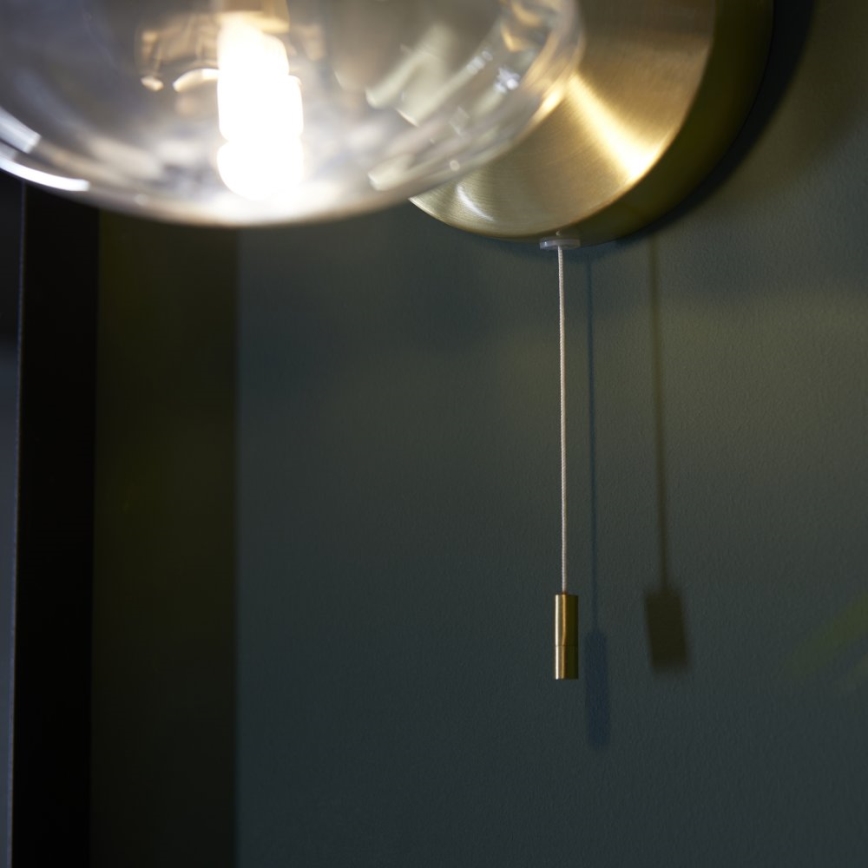 Nástenná lampa so skleneným tienidlom svieti. Z lampy visí šnúrka so zlatým spínačom. Lampa je pripevnená na tmavozelenej stene.