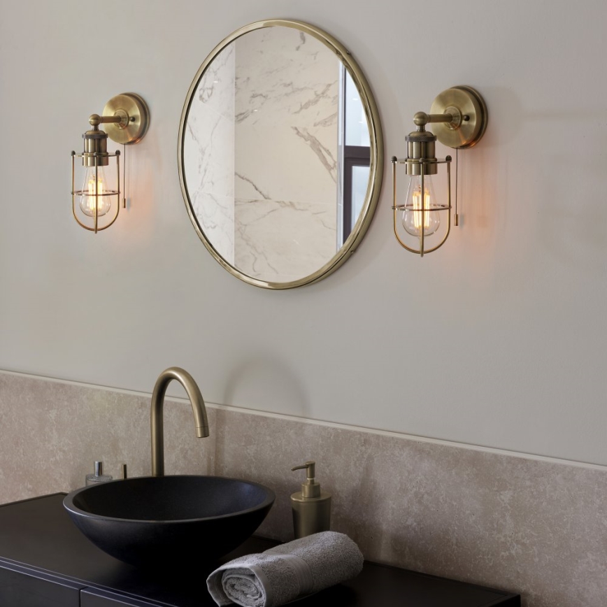 Dve nástenné svietidlá svietia v kúpeľni. Svietidlá sú z mosadze, s kovovými klietkami okolo žiaroviek. Nad umývadlom je okrúhle zrkadlo.