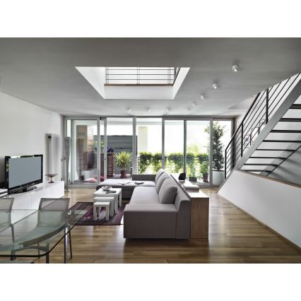 Moderná sedačka v obývacej izbe s drevenou podlahou a veľkými oknami s výhľadom na záhradu. V pozadí sú schody a televízor.