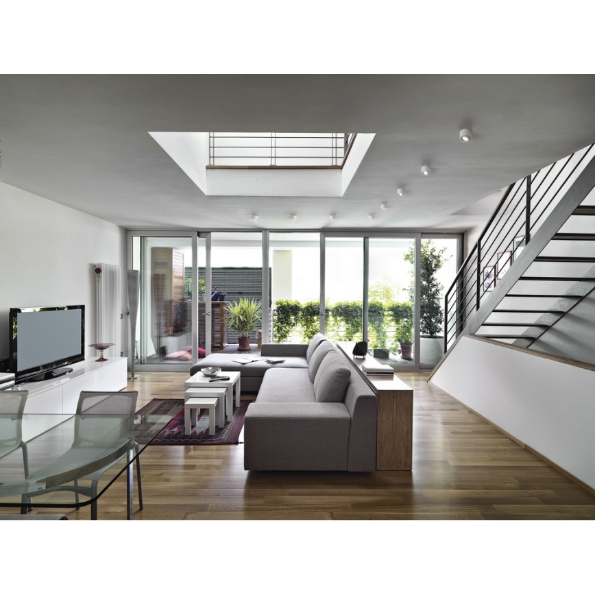 Moderná sedačka v obývacej izbe s drevenou podlahou a veľkými oknami s výhľadom na záhradu. V pozadí sú schody a televízor.