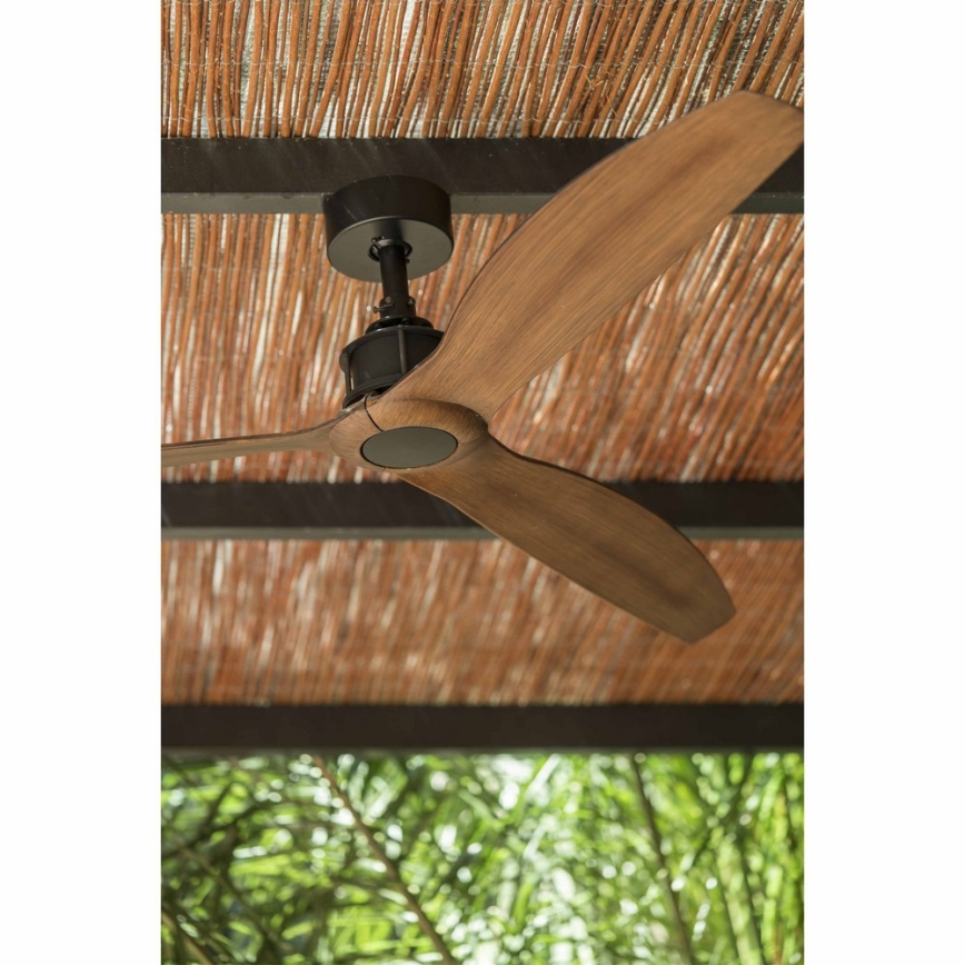 Drevený stropný ventilátor s tmavosivým krytom sa otáča pod hnedou slamkovou strechou. Za strechou je vidieť rozostrené zelené lístie.