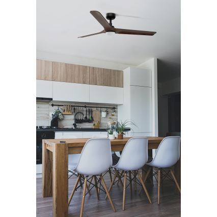 Drevený stropný ventilátor s troma lopatkami sa otáča v kuchyni s dreveným jedálenským stolom a bielymi stoličkami. Kuchyňa má moderný dizajn s drevenými a bielymi skrinkami.