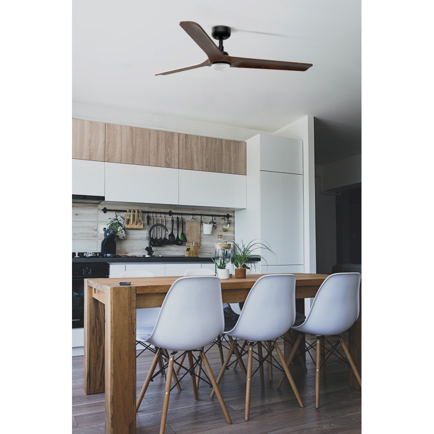 Drevený stropný ventilátor s troma lopatkami sa otáča v kuchyni s dreveným jedálenským stolom a bielymi stoličkami. Kuchyňa má moderný dizajn s drevenými a bielymi skrinkami.