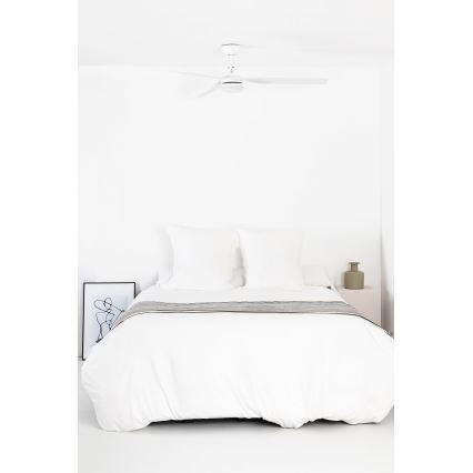 Biela posteľ s bielou prikrývkou a sivým prehozom je umiestnená v bielej miestnosti. Nad posteľou je biely stropný ventilátor. Na nočnom stolíku stojí malá zelená váza.