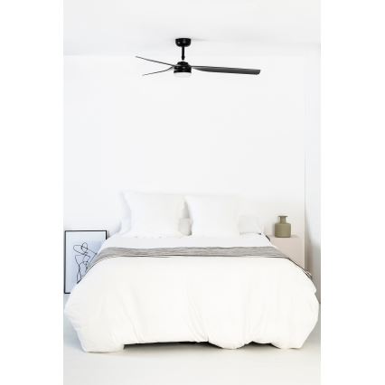 Čierny stropný ventilátor s troma lopatkami rotuje v spálni nad posteľou s bielou posteľnou bielizňou. Vedľa postele je malý nočný stolík s vázičkou.