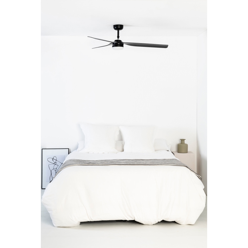 Čierny stropný ventilátor s troma lopatkami rotuje v spálni nad posteľou s bielou posteľnou bielizňou. Vedľa postele je malý nočný stolík s vázičkou.