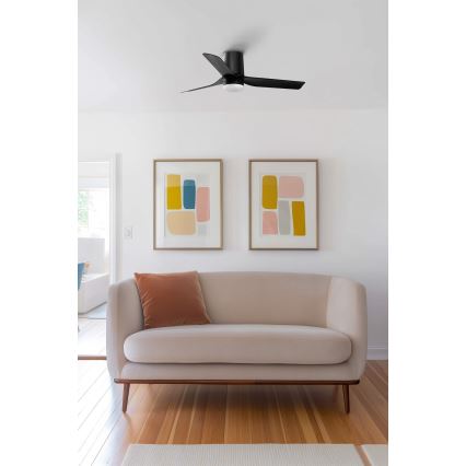 Stropný ventilátor s troma lopatkami, čiernej farby, sa nachádza v obývacej izbe s béžovou sedačkou a dvoma obrazmi s geometrickými vzormi na stene.