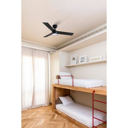 Čierny stropný ventilátor s tromi lopatkami sa otáča v detskej izbe. Izba obsahuje drevené poschodové lôžko s červenými ochrannými prvkami a svetlými posteľnými plachtami. Podlaha je z drevených parkiet.