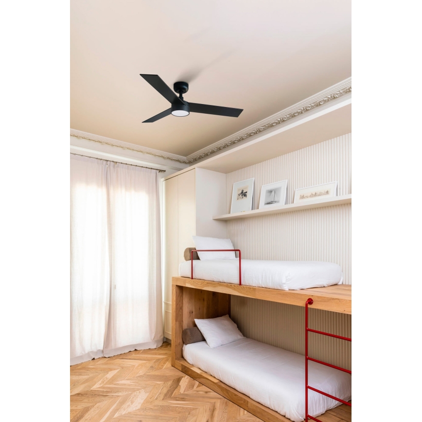 Čierny stropný ventilátor s tromi lopatkami sa otáča v detskej izbe. Izba obsahuje drevené poschodové lôžko s červenými ochrannými prvkami a svetlými posteľnými plachtami. Podlaha je z drevených parkiet.