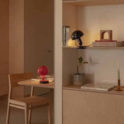 Červená stolová lampa svieti na stole v svetlej drevenej miestnosti. Vedľa nej je drevená stolička. Na poličke v pozadí je čierna stolová lampa a knihy s názvami „100 INTERIORS AROUND THE WORLD“ a „NEUTRA“.