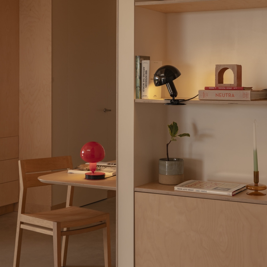 Červená stolová lampa svieti na stole v svetlej drevenej miestnosti. Vedľa nej je drevená stolička. Na poličke v pozadí je čierna stolová lampa a knihy s názvami „100 INTERIORS AROUND THE WORLD“ a „NEUTRA“.