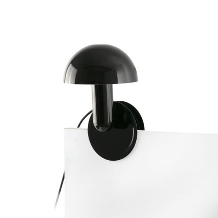 Čierna stolová lampa s kupolovitým tienidlom je pripevnená k bielej ploche. Lampa je upevnená pomocou kruhového klipu. Elektrický kábel je viditeľný.