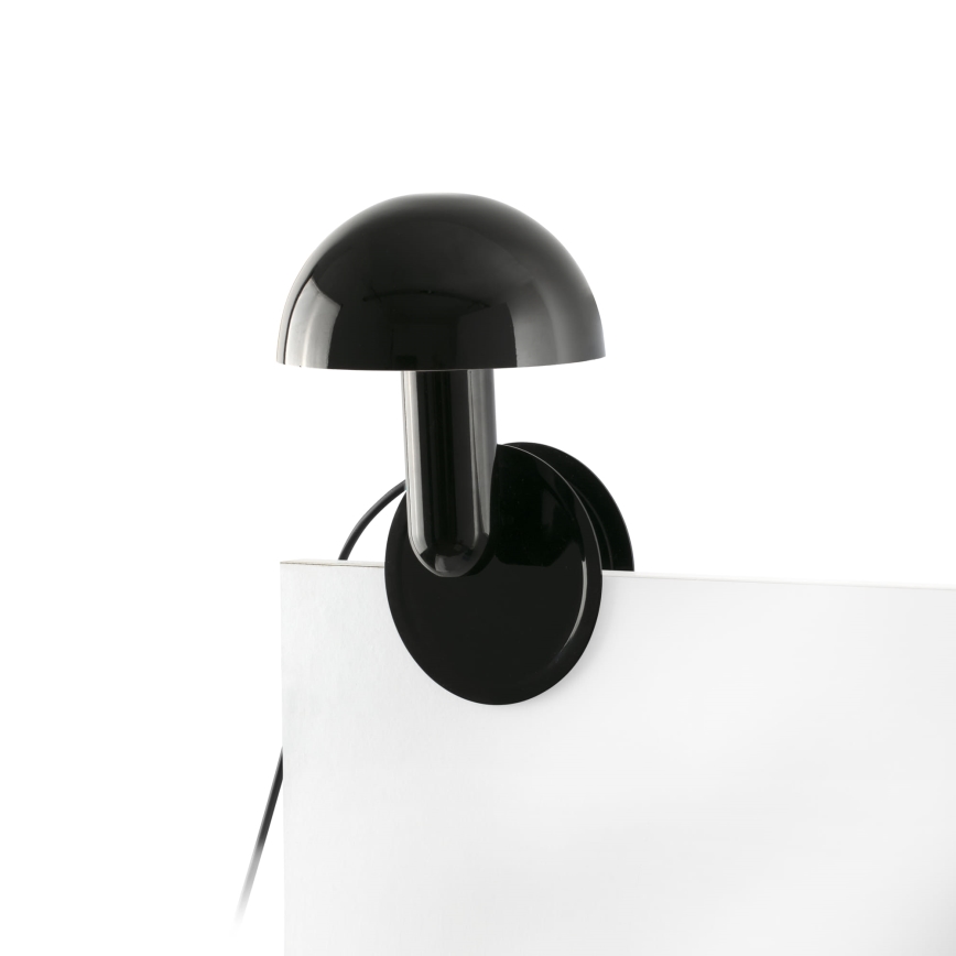 Čierna stolová lampa s kupolovitým tienidlom je pripevnená k bielej ploche. Lampa je upevnená pomocou kruhového klipu. Elektrický kábel je viditeľný.