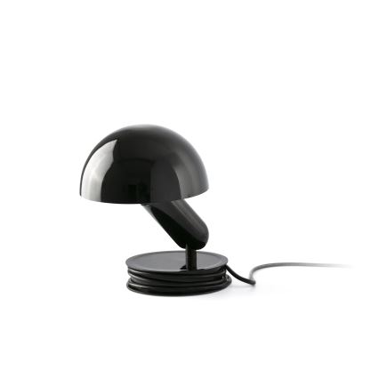 Stolová lampa s čiernym tienidlom v tvare huby osvetľuje priestor. Je umiestnená na čiernej podložke, okolo ktorej je navinutý čierny kábel.