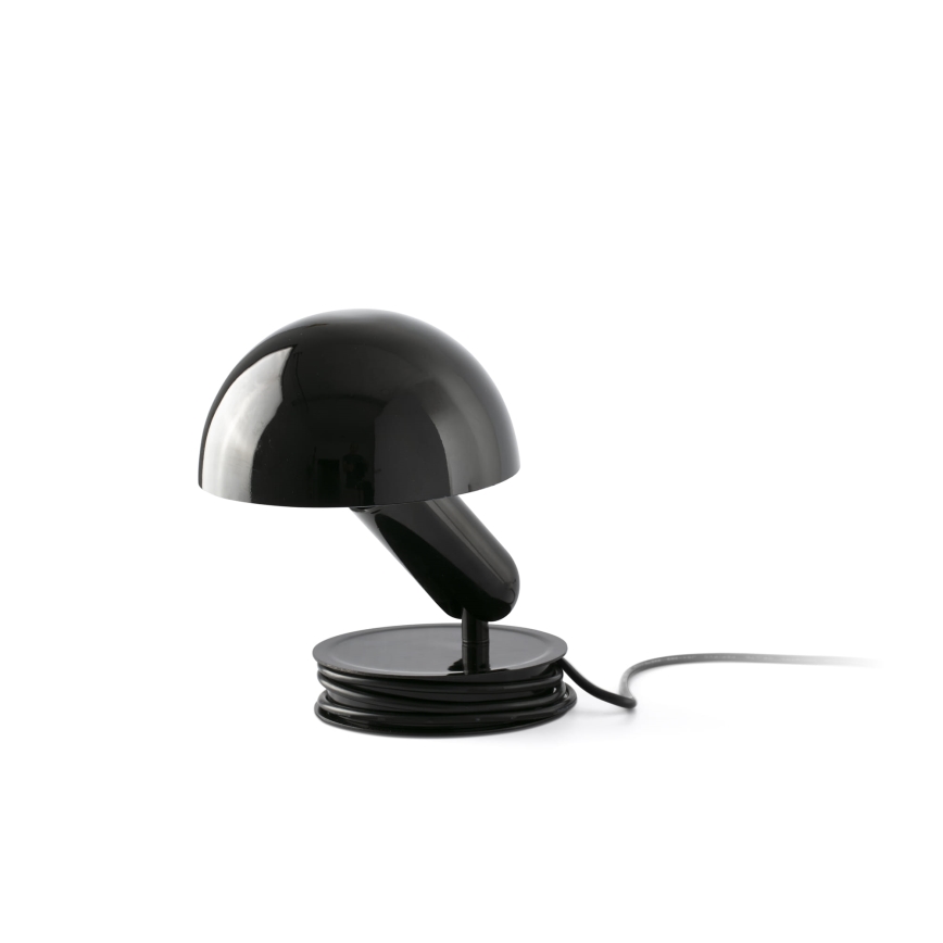 Stolová lampa s čiernym tienidlom v tvare huby osvetľuje priestor. Je umiestnená na čiernej podložke, okolo ktorej je navinutý čierny kábel.