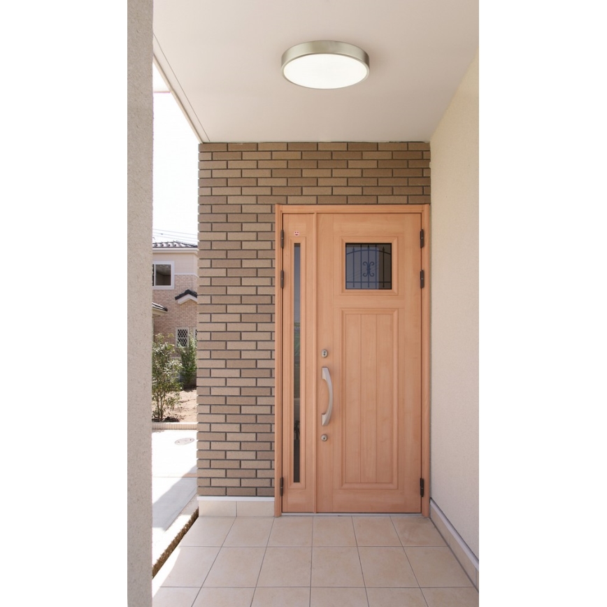 Dvere z svetlého dreva sa nachádzajú v exteriéri domu. Dvere sú osadené v tehlovej stene. Nad dverami je stropné svietidlo.