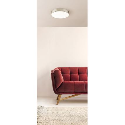 Okrúhle stropné svietidlo je pripevnené k stropu. Pod ním sa nachádza bordová pohovka na drevených nožičkách a svetlohnedý koberec.