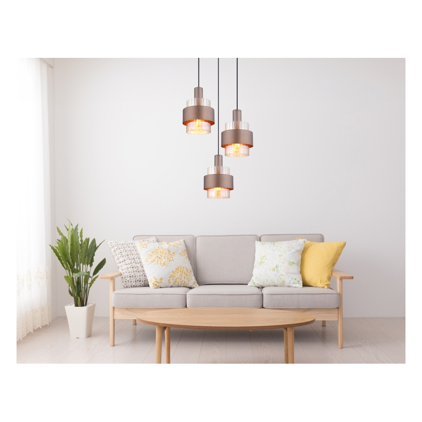 Trojité závesné svietidlo visí nad pohovkou v obývacej izbe. Svietidlá sú kovové, s priehľadnými sklenenými vložkami a teplým osvetlením. Pohovka je svetlošedá a stojí pred nízkym dreveným konferenčným stolíkom.