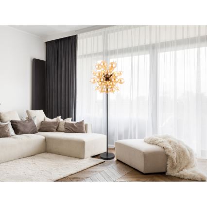 Stojacia lampa s rozsiahlym tienidlom z mnohých sklenených guľôčok svieti v obývacej izbe s rohovou sedačkou a svetlým kobercom. Lampa stojí na tmavosivej podstave. V pozadí sú svetlé závesy a okná.