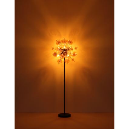 Stojacia lampa svieti. Jej tienidlo pozostáva z mnohých malých, priehľadných sklenených guličiek, ktoré sú usporiadané do guľovitého tvaru. Lampa stojí na tmavo-hnedej podlahe pred hnedou stenou.