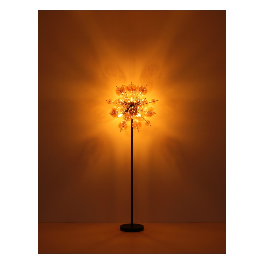 Stojacia lampa svieti. Jej tienidlo pozostáva z mnohých malých, priehľadných sklenených guličiek, ktoré sú usporiadané do guľovitého tvaru. Lampa stojí na tmavo-hnedej podlahe pred hnedou stenou.