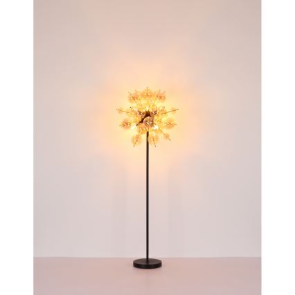 Stojacia lampa svieti. Jej tienidlo tvorí sústava mnohých malých sklenených guľôčok pripevnených k čiernemu kovovému stonku s čiernou podstavou. Lampa stojí na svetlej podlahe pred svetlou stenou.