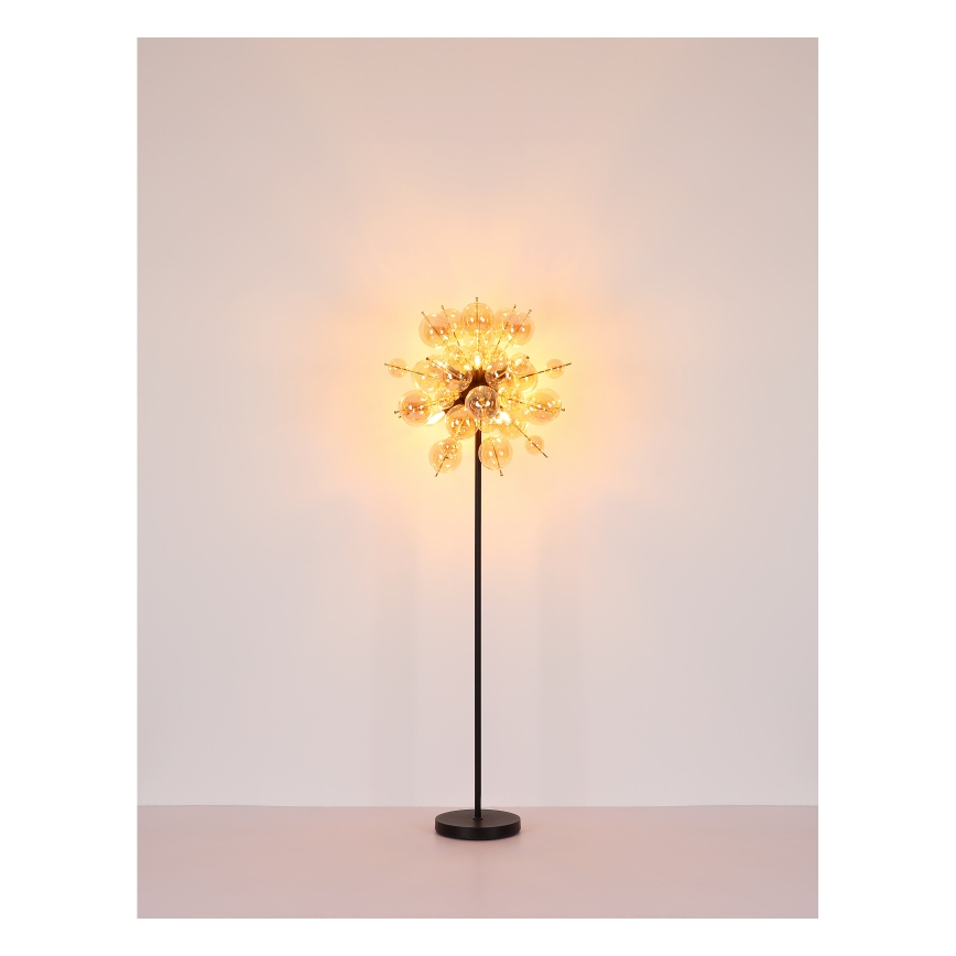 Stojacia lampa svieti. Jej tienidlo tvorí sústava mnohých malých sklenených guľôčok pripevnených k čiernemu kovovému stonku s čiernou podstavou. Lampa stojí na svetlej podlahe pred svetlou stenou.