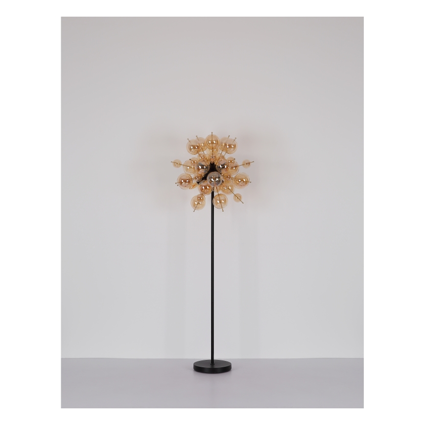 Stojacia lampa s vysokým čiernym stojanom a svietidlom zloženým z viacerých malých jantárových sklenených guľôčok usporiadaných v tvare rozvetveného kvetu pred bielou stenou.