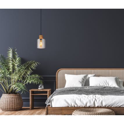 Závesné svetlo visí v spálni. Svetlo je sklenené s dreveným podstavcom. V pozadí je posteľ s ratanovým čelom a rastlina v kvetináči.