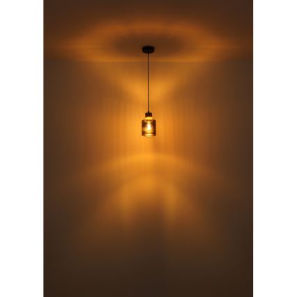 Závesné svietidlo svieti v miestnosti. Svietidlo má valcový sklenený tienidlo a čierny kovový držiak. Svetlo vytvára na stene rozsiahly svetelný kužeľ.