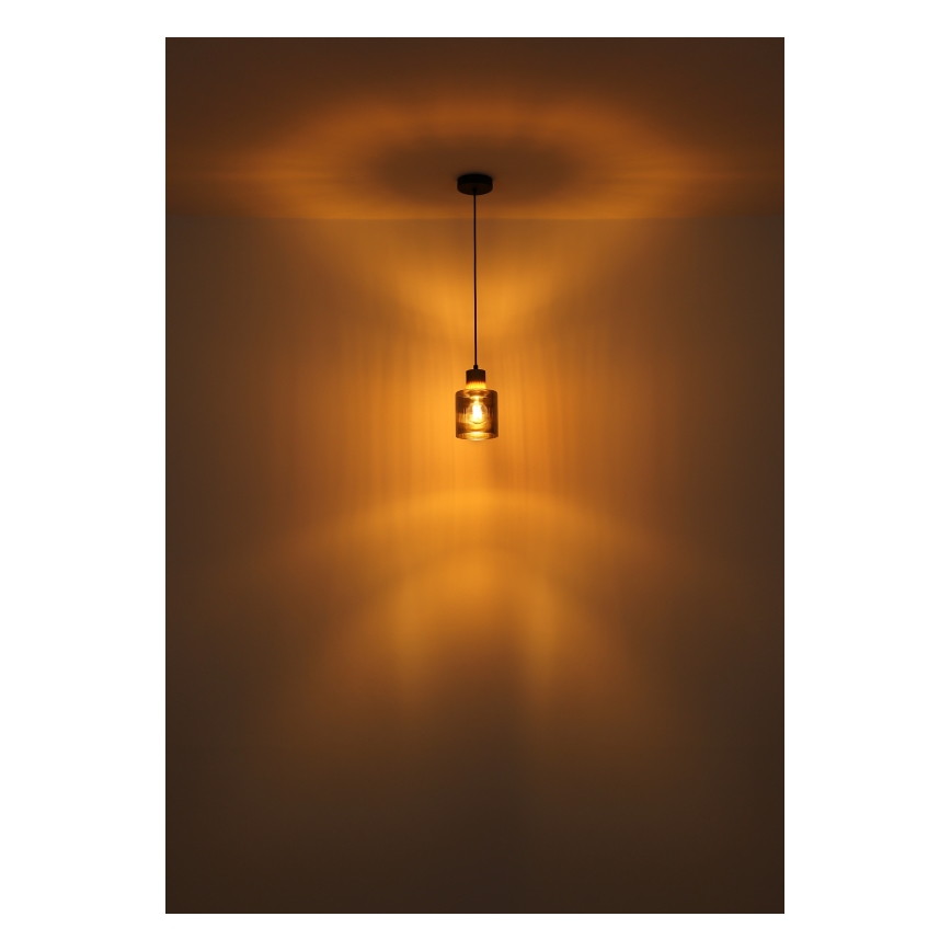 Závesné svietidlo svieti v miestnosti. Svietidlo má valcový sklenený tienidlo a čierny kovový držiak. Svetlo vytvára na stene rozsiahly svetelný kužeľ.