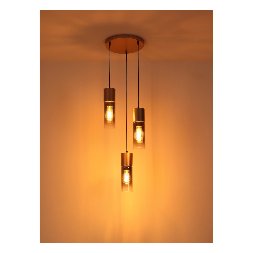 Trojité závesné svietidlo svieti v priestore s oranžovou stenou. Každé svietidlo má valcový tienidlo zo skla a kovový zlatý rám. Svietidlá sú zavesené z kruhového stropného držiaka.