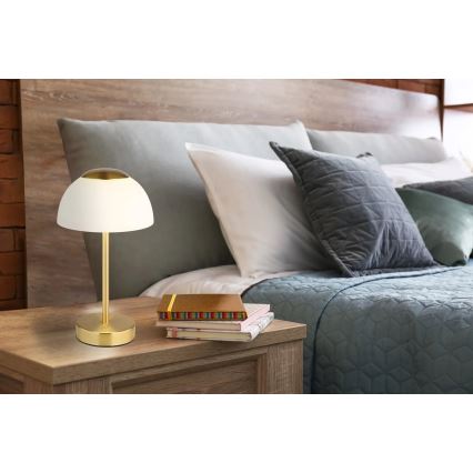 Stolová lampa so zlatým podstavcom a bielym tienidlom v tvare kupoly stojí na nočnom stolíku v spálni. Na stolíku sú uložené knihy. Lampa je umiestnená vedľa postele so sivými a bielymi vankúšmi.