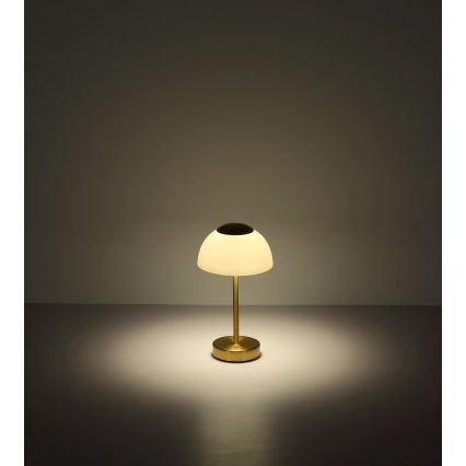 Stolová lampa s polguľovitým tienidlom zo skla svieti. Je zlatá, umiestnená na svetlom povrchu. Pozadí tvorí svetlá stena.