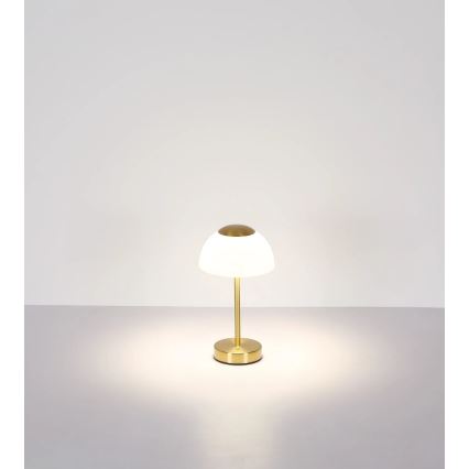 Stolová lampa svieti. Má zlatú kovové podstavu a stonku a biely sklenený tienidlo v tvare kupoly. Stojí na svetlom povrchu pred bielou stenou.