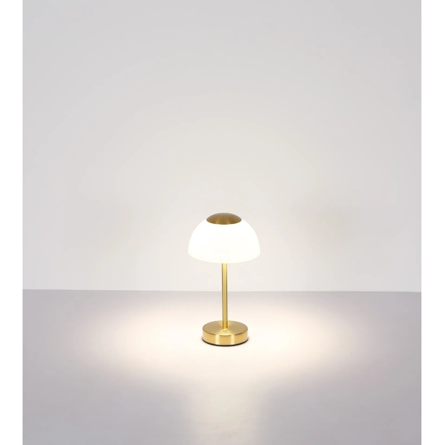 Stolová lampa svieti. Má zlatú kovové podstavu a stonku a biely sklenený tienidlo v tvare kupoly. Stojí na svetlom povrchu pred bielou stenou.