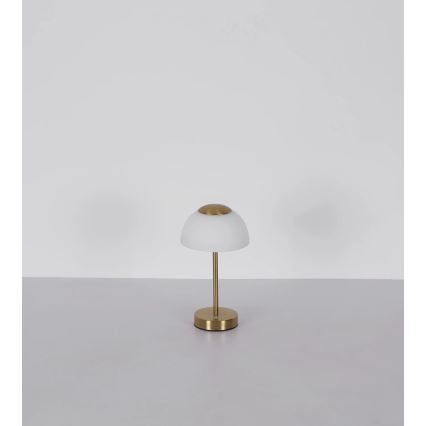 Stolová lampa s bielym tienidlom v tvare kupoly a zlatou kovovou základňou stojí na bielom stole pred bielou stenou.