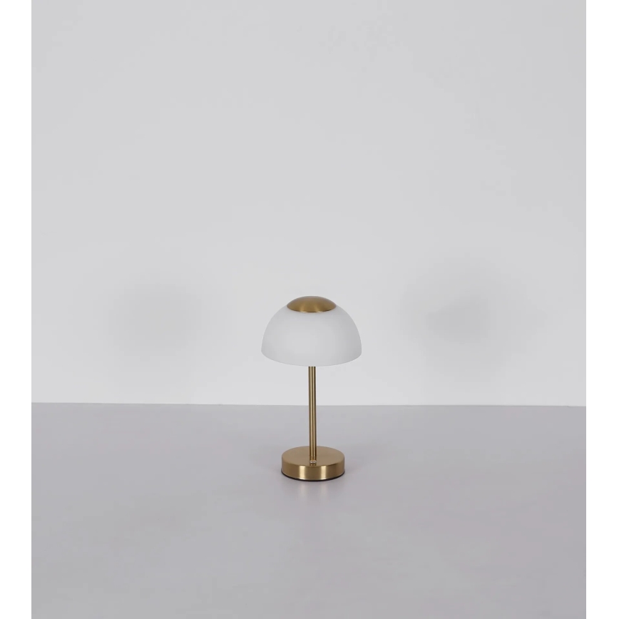 Stolová lampa s bielym tienidlom v tvare kupoly a zlatou kovovou základňou stojí na bielom stole pred bielou stenou.