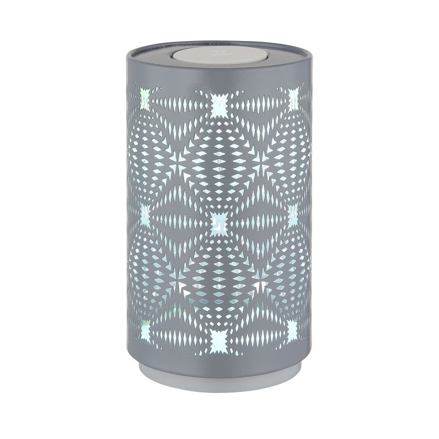 Šedá valcová lampa s vyrezávaným geometrickým vzorom svieti jemným svetlom. Stojí na bielom podstavci na bielom pozadí.