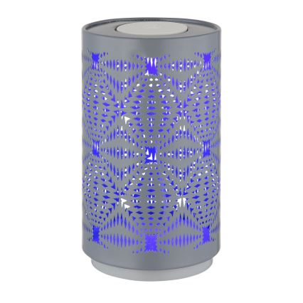 Sivá kovová lampa valcovitého tvaru s vyrezávaným vzorom svieti fialovým svetlom. Lampa stojí na bielom podstavci.